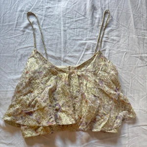 Glittrig blommig croppad linnetopp - Supersöt croppad linnetopp med tunna axelband och volangig passform. Toppens tyg är blommigt med inslag av guldglittriga detaljer i hela mönstret, och färgerna går i ljusa toner av beige, lila och rosa. Perfekt för dig som vill sticka ut med en unik och glittrig look.