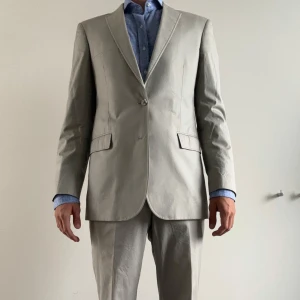 Beige kostym från J.Lindeberg - Snygg beige kostym från J.Lindeberg i klassisk modell med en modern twist. Kavajen har två knappar, fickor med lock och lätt vadderade axlar. Tillverkad i 90 % bomull och 10 % polyester för en lätt och bekväm känsla. Designad med en något sportigare känsla än traditionella kostymer, gjord för golfbanan. Kavaj i storlek 50, byxa i storlek 48. Hör av dig vid frågor 😊