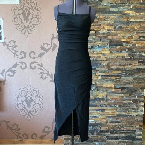 Svart glittrig långklänning New Collection - Supersnygg svart långklänning med glittrigt tyg och smala axelband från New Collection. Vadderade kupor. Klänningen har en asymmetrisk slits framtill och figurnära passform som framhäver siluetten. Uppskattad storlek 38