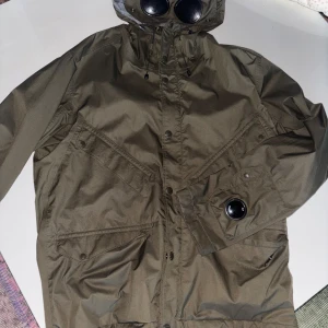 C.P. Company Goggle Jacket olivgrön - Säljer en ikonisk olivgrön Goggle Jacket från C.P. Company. Jackan har huva med inbyggda goggles, stora fickor framtill och är tillverkad i slitstark polyester. Perfekt för dig som vill sticka ut med streetwear-vibe och smarta detaljer.