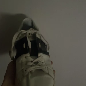 Valentino Rockstud vita sneakers - Snygga vita Valentino Rockstud sneakers med svarta detaljer och ikoniska nitar på sidorna. Skorna har snörning, platt sula och rund tå. Tillverkade i skinn för en lyxig känsla och cool look. Perfekta för dig som vill sticka ut med en exklusiv touch.