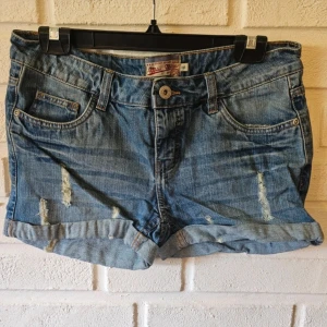 jeansshorts från Miss Milla - Snygga jeansshorts från Miss Milla med slitna detaljer och upprullade benslut. Klassisk femficksmodell med knapp och dragkedja framtill. Perfekta för sommardagar och ger en avslappnad vibe till din outfit.