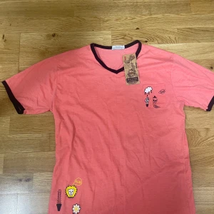 Rosa t-shirt med tryck från Bershka - Rosa t-shirt från Bershka med mörka kontrastkanter vid hals och ärmslut. V-ringad modell med coola tryck som lejon, blomma och tecknade figurer. Perfekt för dig som vill sticka ut lite extra. Skön bomullskänsla och avslappnad passform.