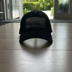 Svart truckerkeps från Hatstore - Svart truckerkeps från Hatstore med mesh på sidorna och baksidan för extra ventilation. Framsidan har ett svartvitt tryck med motiv av arbetare på en balk. Justerbar passform baktill. Perfekt för dig som vill ha en cool och avslappnad stil.