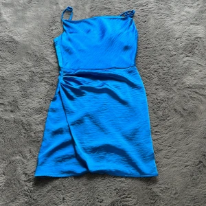 Blå satin klänning Gina Tricot - Supersnygg blå kort klänning från Gina Tricot i glansigt satinliknande material. Klänningen har smala axelband, omlott-detalj vid höften och dragkedja i ryggen. Perfekt för dig som vill sticka ut med en poppig färg och enkel, stilren design.