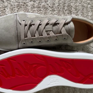 Beige mockasneakers från Louboutin - Snygga sneakers från Christian Louboutin i beige mocka med klassisk röd sula och vita kanter. Skorna har rund tå, snörning och lyxig känsla med läderfoder. Perfekta för dig som vill sticka ut med exklusiva detaljer och stilren design.