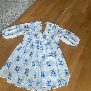 Vit blommig kortklänning med v-ringning - Superfin vit kortklänning med blått blommönster och v-ringning. Klänningen har långa ärmar och en lätt rynkad midja som ger en luftig känsla. Perfekt för varma dagar och har en romantisk vibe med sina blå detaljer.