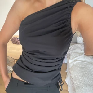 Svart oneshoulder topp från Zara - Säljer en svart oneshoulder topp från Zara. Toppen har draperad sida och snörning för en snygg passform. Gjord i mjukt, stretchigt material som sitter tight och markerar axeln. 🖤 använd 1 gång. Svinsnygg
