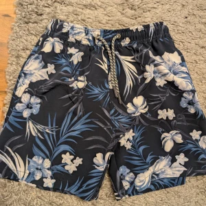 Blå blommiga badshorts från D.M.D stl 146-152 - Snygga badshorts från D.M.D med blått och vitt blommönster och tropiska blad. Shortsen har elastisk midja med snörning och innerbyxa i mesh för extra komfort. Perfekta för strand eller poolhäng i sommar!