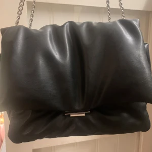 Svart axelväska i skinn från H&M - Säljer en svart axelväska från H&M i mjukt skinn med puffig design. Väskan har en silverfärgad kedjerem och ett stilrent metallspänne framtill. Insidan har dragkedjefack för småsaker. Perfekt för dig som gillar en trendig och clean look.