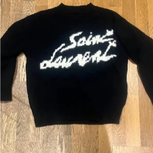 Svart stickad Saint Laurent tröja - Svart stickad tröja från Saint Laurent med vit, stor logga framtill i coolt grafiskt mönster. Klassisk rund hals och långa ärmar. Perfekt för dig som vill ha en statement piece med lyxig känsla.