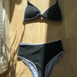 Svart bikini med vita detaljer - Snygg svart bikini med höga trosor och trekants-bh. Bikinin har vita sicksack-detaljer längs kanterna och ribbad struktur i tyget. Perfekt för stranden eller poolen i sommar.