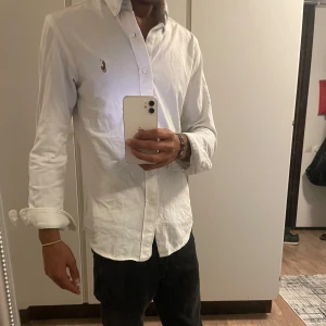 Vit skjorta från Ralph Lauren - Klassisk vit skjorta från Ralph Lauren med broderad logga på bröstet. Skjortan har lång ärm, knappar framtill och en snygg krage. Perfekt för dig som gillar stilrena och tidlösa plagg. Passformen är normal och materialet känns mjukt och bekvämt.