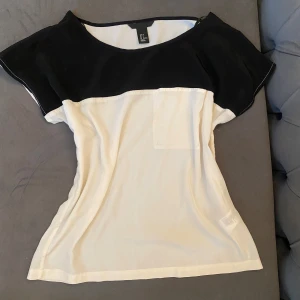 Svartvit blus med kort ärm - Snygg svartvit blus från H&M i storlek 34 med kontrastfärgade partier. Överdelen och ärmarna är svarta medan resten av blusen är vit. Modellen har korta, lösa ärmar och en liten bröstficka framtill. Perfekt för en stilren och enkel look💗