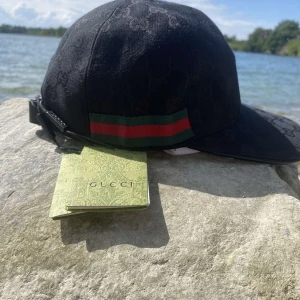 Svart keps från Gucci med GG-mönster - Snygg svart keps från Gucci med diskret GG-monogram över hela kepsen och klassiska gröna och röda ränder på sidan. Justerbar rem bak och tillverkad i bomull. Kepsen kommer med originalpåse och tags.