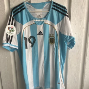 Argentina Messi 19 VM 2006 tröja Adidas - Säljer en klassisk Argentina fotbollströja från VM 2006 med Messi och nummer 19 på ryggen. Tröjan är randig i ljusblått och vitt, har svarta Adidas-ränder på axlarna och officiellt AFA-märke på bröstet. Tillverkad i lätt och ventilerande material.