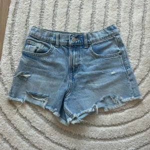 Ljusblå jeansshorts från Zara - Snygga ljusblå jeansshorts från Zara med råa kanter och slitna detaljer. Klassisk femficksmodell med bälteshällor och knappstängning. Perfekta för varma dagar och ger en avslappnad vibe.