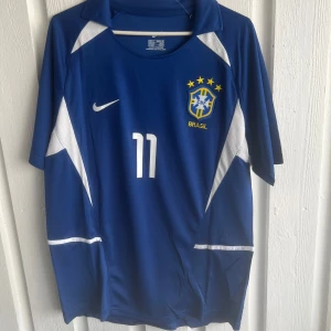 Blå Brasil Ronaldinho fotbollströja Nike - Snygg blå Brasilien fotbollströja från Nike med Ronaldinho och nummer 11 på ryggen. Tröjan har vita detaljer, korta ärmar och klassisk krage. Tillverkad i lätt och ventilerande material, perfekt för match eller träning.
