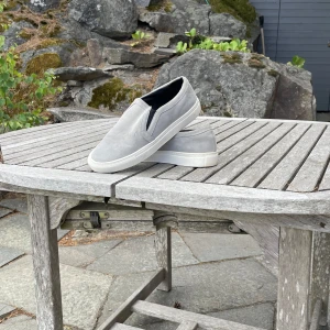 Grå slip-on sneakers i mocka, sandro - Stilrena grå slip-on sneakers med vit sula och ovandel i mjuk mocka. Skorna har elastiska partier på sidorna för enkel påtagning och en clean look. Perfekta för dig som gillar stilren stil och vill ha något som funkar till det mesta. Märket heter sandro och dessa skor har jag använt väldigt lite eftersom att de blev för små väldigt fort. Nypris cirka 3000kr 😊