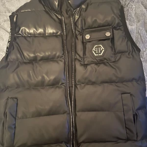 Svart dunväst från Philipp Plein - Snygg svart dunväst från Philipp Plein med quiltad design och dragkedja framtill. Västen har en bröstficka med lock och logga, samt två sidofickor. Tillverkad i glansig polyester som ger en exklusiv känsla. Har använt den 3-4 gånger. Köpte den för 2 år sen. Vid snabbaffär kan jag gå ner i pris
