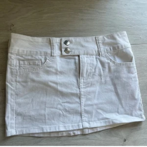 Vit jeanskjol med fickor - Snygg vit kort jeanskjol med klassiska fickor fram och dubbla silverknappar i midjan. Kjolen har bälteshällor och en stilren, rak passform. Perfekt för dig som gillar en clean och fräsch look.