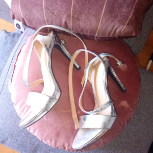 Silvriga klackpumps från Zara - Snygga silvriga pumps från Zara med hög klack och smala remmar. Skorna har öppen tå och justerbart spänne runt vristen. Perfekta för dig som vill sticka ut med metallic-look och elegant design.