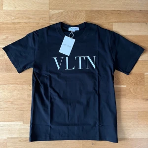 Svart VLTN t-shirt från Valentino - Svart t-shirt från Valentino med stor vit VLTN-logga på bröstet. Klassisk rund halsringning och korta ärmar. Tillverkad i mjuk bomull för en skön känsla och clean look. Perfekt för dig som gillar stilrena designer och exklusiva märken.