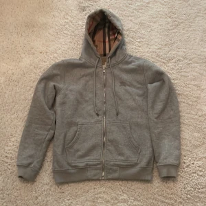 Grå hoodie från Burberry med dragkedja - Säljer en grå hoodie från Burberry i bomull med klassiskt rutigt foder i huvan och längs insidan. Tröjan har dragkedja, snörning i huvan, två stora fickor fram och broderad Burberry-logga på bröstet. Perfekt för dig som gillar stilren design med lyxig touch. Kan sänkas vid snabb affär!!!! Vill få bort den så snabbt som möjligt!!!