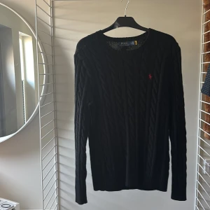 Svart tröja Polo Ralph Lauren - Svart kabelstickad tröja från Polo Ralph Lauren med klassisk röd broderad logga på bröstet. Tröjan har rund halsringning, lång ärm och ribbade muddar. Perfekt för dig som gillar en stilren och tidlös look. Storlek S men passar M då den är stor i storlek.