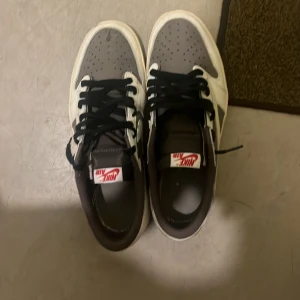 Nike Air Jordan 1 Low Travis Scott - Nike Air Jordan 1 Low Travis Scott med omvänd Swoosh i vitt skinn, grå mocka och svarta skosnören. Röd Air Jordan-logga och Cactus Jack-broderi på hälen. Snygg kontrast mellan de neutrala färgerna och detaljerna gör dem riktigt unika.