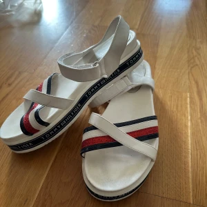Vita sandaler Tommy Hilfiger - Snygga vita sandaler från Tommy Hilfiger med breda remmar och coola röda och marinblå glitterdetaljer. Sulan har logotypen runt om och sandalerna har kardborreknäppning för enkel påtagning. Perfekta för sommaren och ger en sportig vibe.