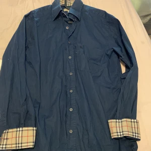 Blå Burberry skjorta med rutiga detaljer - Snygg blå skjorta från Burberry i klassisk passform. Skjortan har långa ärmar, knappar framtill och ikoniska rutiga detaljer på insidan av manschetterna och kragen. Tillverkad i bomull med lite stretch för extra komfort. Perfekt för dig som gillar stilrena plagg med en twist. Ostyrkt men hinner nog stryka innan du köper. Liten skada vid fickan men har limmat och det syns knappt