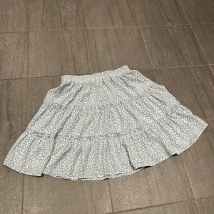 Blå volangkjol med vitt mönster  - Supersöt kort volangkjol i ljusblått med vitt mönster. Kjolen har resår i midjan och flera lager volanger. 