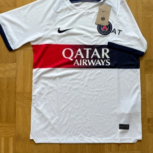 PSG vit matchtröja Nike M - Paris Saint-Germain fotbollströja från Nike i vitt med bred röd och marinblå rand över bröstet och Qatar Airways-tryck. PSG-emblem på bröstet, Dri-FIT-material som andas och korta ärmar med marinblå detaljer. Perfekt för dig som älskar fotboll och vill ha en snygg supportertröja.