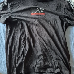 Svart EA7 Emporio Armani t-shirt - Svart t-shirt från EA7 Emporio Armani med tryckt logga på bröstet och axlarna. Klassisk passform och korta ärmar. Tillverkad i mjuk bomull som känns skön mot huden. Perfekt för dig som gillar stilrena och exklusiva plagg med tydlig logga.
