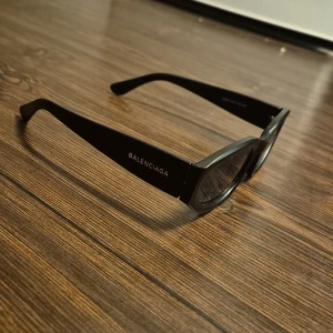 Svarta Balenciaga solglasögon - Snygga svarta solglasögon från Balenciaga med rektangulära bågar och diskret logga på sidan. Glasögonen har en stilren och modern look med breda skalmar och mörka linser. Perfekta för dig som vill ha en clean och exklusiv vibe.