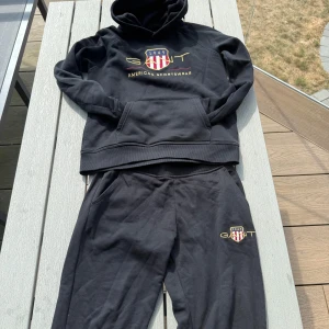 Svart hoodie från GANT med brodyr - Svart hoodie från GANT med broderad logga och texten 'American Sportswear' på bröstet. Tröjan har huva, känguruficka och ribbade muddar. Materialet är mjuk bomull, perfekt för chill dagar. Klassisk och sportig look med snygga detaljer i rött, vitt och guld. Sett från gant säljer båda för 500kr