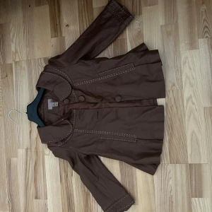 Trekvarts ärmad jacka från H&M  - Snygg brun trekvarts ärmad jacka från H&M med stora klädda knappar och dekorativa vita sömmar längs krage, fickor och ärmslut. Jackan har en klassisk form med bred krage och lång ärm, perfekt för dig som gillar retrovibbar och stilrena detaljer.