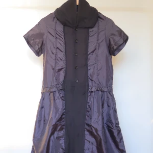 Svart kortklänning Comme des Garçons - Unik svart kortklänning från Comme des Garçons med glansig finish, korta ärmar och hög krage. Klänningen har knappar framtill och markerad midja. Materialet känns lyxigt och har en lätt struktur, perfekt för dig som gillar statement-plagg.