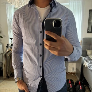 Randig skjorta från Savvy Citizen S - Snygg skjorta från Savvy Citizen i storlek S. Skjortan är vit med tunna blå ränder, har klassisk krage och svarta knappar. Tillverkad i bomull och har lång ärm. Perfekt för dig som gillar stilrena och fräscha plagg med en modern touch.