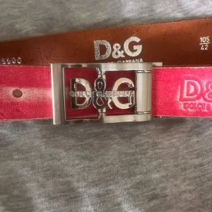 Rosa D&G skärp i skinn med silverdetaljer - Snyggt rosa bälte från Dolce & Gabbana i äkta skinn med stor silverfärgad D&G-logga på spännet. Bältet har präglad logga längs remmen. Perfekt accessoar för att lyfta din outfit.