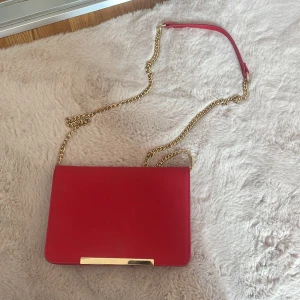 Röd kuvertväska med guldkedja - Snygg röd kuvertväska med stilren design och guldfärgad kedjerem. Väskan har en slät yta och en elegant gulddetalj framtill. 