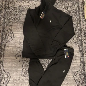 Svart ralph lauren tracksuit - Svart tracksuit från Polo Ralph Lauren med klassisk huva och snörning. Hoodien har en broderad vit logga på bröstet. Mjuk och skön bomullskvalitet, perfekt för chill dagar. Matchande svarta byxor med dragsko och logga ingår. Storlek M men kan passa S också. Vid frågor eller info kom Dm. Pris kan diskuteras vid snabbaffär!