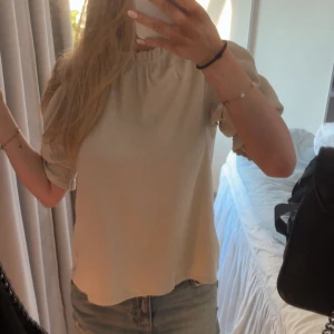 Beige blus med puffärm och volang - Supersöt beige blus med lagomt puffiga korta ärmar och volangdetalj vid halsringningen. Blusen har en lätt skrynklig struktur och stängs med en liten knapp i nacken. Perfekt att styla med jeans eller en kjol! Endast använd ett fåtal gånger. 🌸