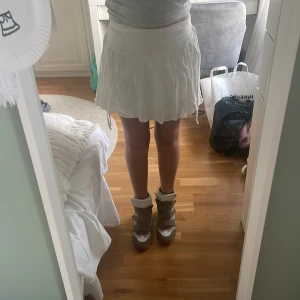 Vit kortkjol med volang och knyt - Supersöt vit kortkjol med volangkant och knyt i sidorna. Kjolen har en bred midjeresår och är gjord i ett lätt, skrynkligt bomullstyg som ger en chill och somrig vibe. Perfekt för varma dagar när du vill känna dig både bekväm och snygg.