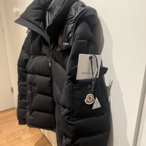 Moncler Jacka - Säljer en svart dunjacka från Moncler med huva och klassisk quiltad design. Jackan har dragkedja framtill, praktiska fickor och ikoniska Moncler-detaljer på ärmen. Perfekt för kalla dagar och riktigt snygg till streetwear-stilen.