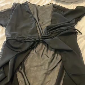 Svart omlottklänning i mesh - Snygg svart omlottklänning i tunn mesh med knyt i midjan. Klänningen har v-ringning och är genomskinlig, perfekt för lager på lager eller över badkläder. Stilren och enkel att styla till olika looks.