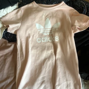 Ljusrosa Adidas t-shirt med logga - Snygg ljusrosa t-shirt från Adidas med stor vit Trefoil-logga framtill. Klassisk rund hals och korta ärmar. Tillverkad i ett sportigt, lätt strukturerat material som känns skönt mot huden. Perfekt för en avslappnad streetstyle-look.