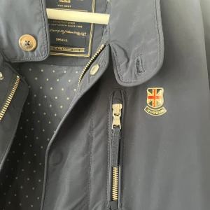 Morris marinblå vindjacka med detaljer - Snygg marinblå vindjacka från Morris med guldfärgade dragkedjor och knappar. Insidan har prickigt foder och jackan har en klassisk badge på bröstet. Perfekt för dig som gillar stilrena och sportiga vibbar. Flera fickor och justerbara detaljer bak.