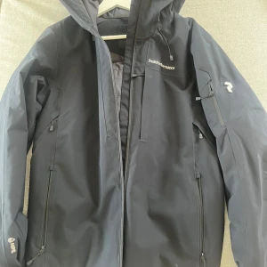 Svart Peak Performance vinterjacka - Peak Performance svart vinterjacka med huva och flera dragkedjeförsedda fickor. Jackan är vadderad och har HIPE-material som skyddar mot väder och vind. Perfekt för kalla dagar och har en clean, sportig look med logga på bröstet och ärmen. Inga skador på jackan. 